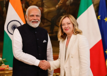Image Prompt: Indian Prime Minister Narendra Modi and Italian Prime Minister Giorgia Meloni shaking hands warmly in an elegant diplomatic hall in Rome, both smiling with confidence, Indian and Italian flags visible in background, Melody toffee pack subtly visible on a table beside them, professional diplomatic photography, cinematic lighting with warm golden tones, photorealistic, 8k resolution, high detail, official state visit atmosphere, --ar 16:9 (1200 x 675 pixels HD size) --no text