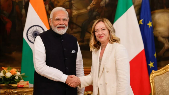 Image Prompt: Indian Prime Minister Narendra Modi and Italian Prime Minister Giorgia Meloni shaking hands warmly in an elegant diplomatic hall in Rome, both smiling with confidence, Indian and Italian flags visible in background, Melody toffee pack subtly visible on a table beside them, professional diplomatic photography, cinematic lighting with warm golden tones, photorealistic, 8k resolution, high detail, official state visit atmosphere, --ar 16:9 (1200 x 675 pixels HD size) --no text